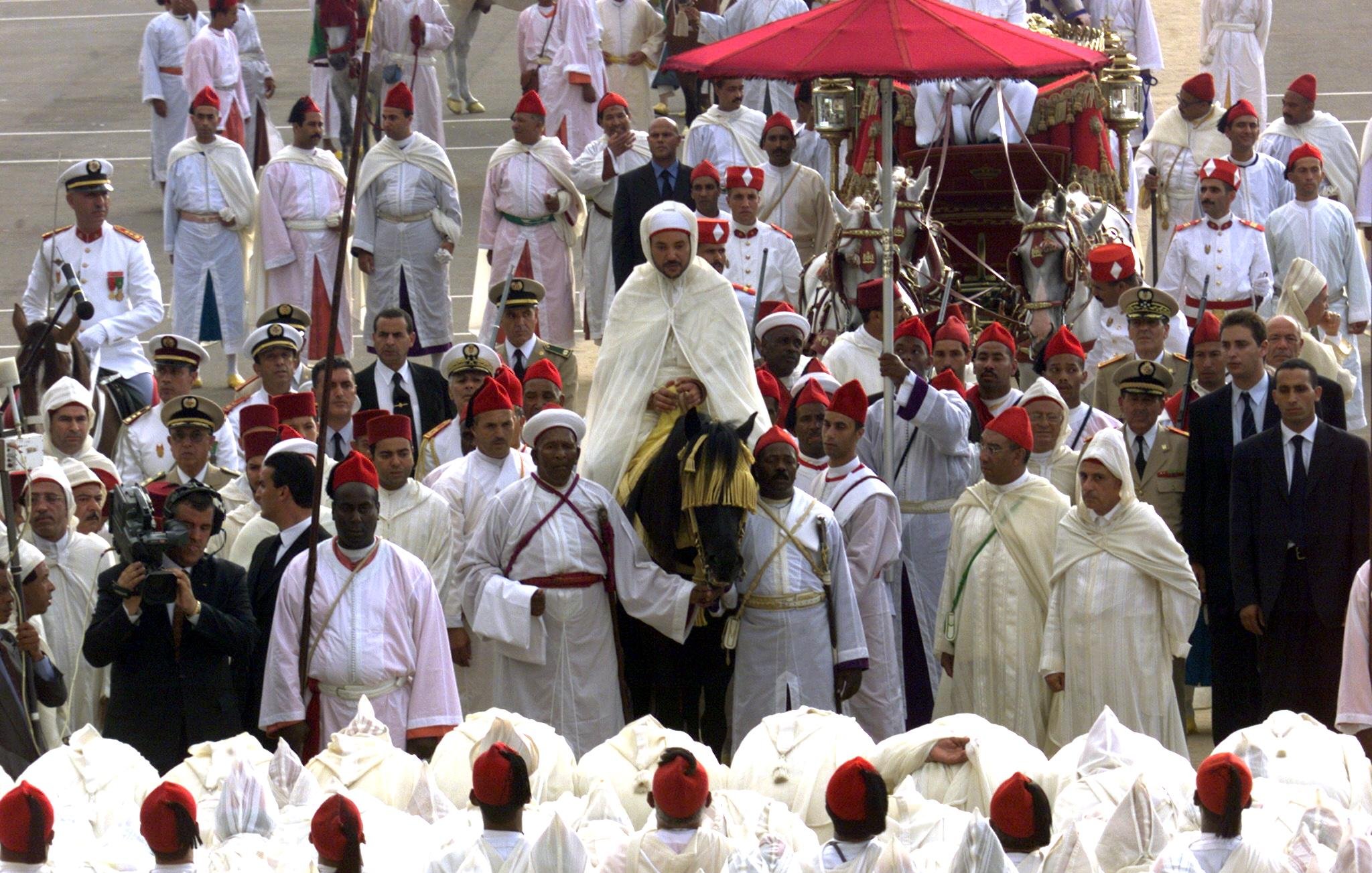 L'image montre un rassemblement cérémonial avec un grand nombre de personnes en tenue traditionnelle. Au centre, une figure importante est portée sur une plate-forme décorée, probablement un leader ou une personnalité éminente. Les participants portent des habits blancs et rouges, indiquant un événement culturel ou religieux. Des officiers en uniformes militaires se trouvent également parmi la foule, et certains photographient la scène. L'ambiance semble festive et respectueuse, typique d'une célébration ou d'une cérémonie officielle.