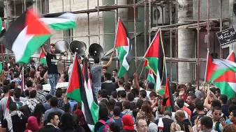 L'image montre une foule rassemblée, brandissant des drapeaux palestiniens. On peut voir des personnes de tous âges, certaines portant des keffiehs, symboles de la culture palestinienne. En arrière-plan, il y a des structures en construction, suggérant un environnement urbain en évolution. L'atmosphère semble dynamique et engagée, avec des expressions de solidarité et de détermination parmi les manifestants. Les haut-parleurs évoquent l'idée que des discours ou des chants sont en cours.