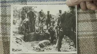 L'image montre un groupe de personnes en uniforme militaire, probablement des soldats, rassemblés autour d'un trou dans le sol. Certains semblent être en train de creuser ou d'examiner le terrain, tandis que d'autres observent. L'ambiance est sérieuse, indicative d'une situation d'urgence ou de conflit. Le tout est en noir et blanc.