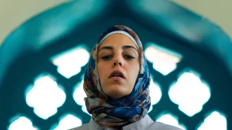 L'image montre une femme portant un hijab coloré, se tenant dans un cadre architectural. La lumière met en valeur son visage et le fond présente des motifs géométriques typiques des designs islamiques, avec des arches et des vitraux. Son expression est sérieuse, ce qui ajoute à la profondeur de la scène.