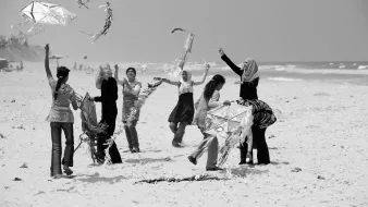 L'image montre un groupe de femmes sur une plage, s'amusant à faire voler des cerfs-volants. Certaines d'entre elles sont en train de tenir ou de manipuler des cerfs-volants, tandis que d'autres semblent rire et profiter du moment. L'environnement est ensoleillé, avec des vagues en arrière-plan. La scène dégage une atmosphère joyeuse et festive, mettant en avant des interactions sociales et des activités de plein air.