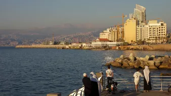 L'image montre un paysage côtier au bord de la mer. On peut voir une promenade avec des personnes qui se tiennent près de l'eau, probablement en train de profiter du coucher de soleil. Au fond, il y a des bâtiments modernes et quelques grues de construction, suggérant un développement urbain. La mer est calme et reflète la lumière dorée du soleil. La scène dégage une atmosphère paisible et agréable.