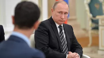 L'image montre un homme assis en costume noir avec une cravate, ayant une expression sérieuse sur le visage. Il semble être en train d'écouter ou de réfléchir pendant une discussion. On peut apercevoir une autre personne au premier plan, de dos, qui ne peut pas être identifiée. L'environnement semble être un bureau officiel, avec des éléments décoratifs typiques d'un cadre institutionnel.