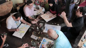 L'image montre un groupe de personnes assises sur le sol dans un espace créatif. Elles semblent engagées dans une activité artistique, utilisant des pinceaux et de la peinture sur des feuilles de papier. Certains participants semblent discuter ou partager des idées, tandis que d'autres se concentrent sur leur travail. L'environnement est coloré, avec des œuvres d'art visibles sur les murs et une palette de peinture au centre. L'ambiance paraît conviviale et collaborative.