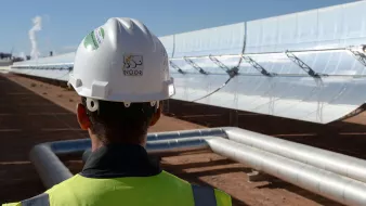 L'image montre un homme portant un casque de sécurité et un gilet de chantier, tourné vers un champ de miroirs solaires. Les miroirs sont orientés vers le soleil et semblent faire partie d'une installation de production d'énergie solaire. À l'arrière-plan, on peut apercevoir des tuyaux métalliques qui transportent probablement un fluide caloporteur. L'environnement est désertique, avec un ciel bleu dégagé qui suggère des conditions solaires idéales.