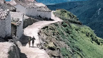 L'image montre un paysage rural avec un sentier serpentant le long d'une colline. De chaque côté du chemin, on peut voir des maisons blanches avec des toits en tuiles. Deux personnes marchent sur le sentier, profitant de la vue. En arrière-plan, des montagnes verdoyantes s'élèvent, créant une atmosphère paisible et naturelle. Le ciel est clair et contribue à l'ambiance sereine de la scène.