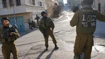 L'image montre des soldats armés en tenue militaire dans une rue apparemment urbaine. Deux d'entre eux sont en position de vigilance, tandis qu'un autre semble communiquer ou surveiller. L'environnement semble être un quartier situé dans une ville, avec des bâtiments en arrière-plan. L'atmosphère pourrait suggérer une situation de tension ou de surveillance.