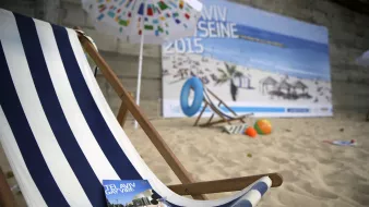 L'image montre une scène de plage avec un transat en bois rayé. Il y a des brochures posées sur le transat, portant des titres promouvant Tel Aviv et ses attractions. En arrière-plan, on peut voir un grand panneau affichant une ambiance estivale, suggérant une plage animée. Le sol est recouvert de sable, et on distingue également des objets colorés comme des ballons. Des parasols, ainsi qu'une vue sur la mer, ajoutent une atmosphère estivale à l'ensemble.