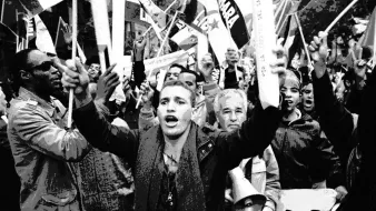 L'image montre une foule en pleine manifestation. On peut voir des personnes brandissant des drapeaux et des pancartes, exprimant leurs opinions avec passion. Au premier plan, un jeune homme s'écrie, tandis que d'autres personnes autour de lui semblent également mobilisées et engagées. L'ambiance générale dégage une forte énergie et un sentiment de solidarité parmi les manifestants. Les visages reflètent une gamme d'émotions, allant de la détermination à l'enthousiasme.