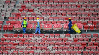L'image montre des travailleurs dans un stade vide. Ils portent des casques de protection et des gilets réfléchissants. Les sièges du stade sont recouverts de plastique, et les travailleurs semblent être en train de nettoyer ou de préparer les lieux. On peut voir des rangées de sièges rouges. L'atmosphère paraît calme et organisée.