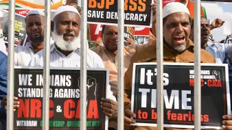 L'image montre un groupe de manifestants qui se tiennent derrière des barreaux. Ils tiennent des pancartes exprimant leur opposition à certaines lois et politiques en Inde, notamment le BJP, le NRC et le CAA. Les manifestants ont des expressions déterminées et sont clairement engagés dans leur cause, appelant à la démocratie et à la résistance. Les pancartes portent des messages en anglais et en d'autres langues, soulignant leur solidarité et leur demande de justice.