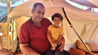 L'image montre un homme souriant assis à l'intérieur d'une tente, tenant un jeune enfant sur ses genoux. L'homme porte une chemise rouge et a l'air détendu et joyeux. L'enfant, qui porte un t-shirt jaune, sourit également et semble heureux. L'intérieur de la tente est simple, avec des coussins au sol, créant une atmosphère conviviale et chaleureuse.