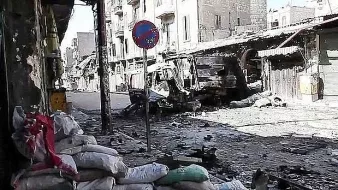 L'image montre une rue dévastée, probablement dans une zone de conflit. On peut voir des bâtiments endommagés et détruits, avec des débris éparpillés sur le sol. Des sacs de sable sont accumulés sur le côté, ce qui suggère des efforts de protection. Un panneau de signalisation est visible, mais l'environnement semble plutôt déserté et en ruines. L'atmosphère dégage un sentiment de désolation et de destruction.