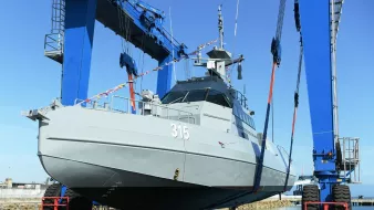 L'image montre un navire militaire en cours de manutention. Il est positionné sur un quai, soutenu par des grues bleues qui le soulèvent avec des sangles. Le bateau est de couleur grise et porte le numéro 315. Le ciel est clair, suggérant une journée ensoleillée. Cette scène évoque des opérations de maintenance ou de construction navale.