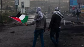 L'image montre deux hommes marchant sur une route, chacun portant un keffieh. L'un d'eux tient un drapeau palestinien, qui est composé de bandes noire, blanche, verte et rouge. L'arrière-plan présente des panneaux publicitaires, des débris et une ambiance brumeuse. Les hommes semblent se diriger vers une direction, symbolisant probablement une manifestation ou une action de revendication.