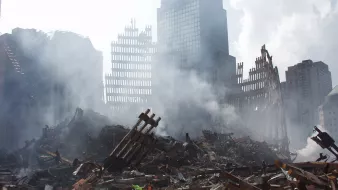 L'image montre un paysage urbain dévasté, avec des débris de bâtiments effondrés et des structures en ruines. De la fumée s'élève dans l'air, créant une atmosphère sombre et chaotique. On peut voir des restes de structures métalliques, avec des morceaux de béton éparpillés sur le sol. L'arrière-plan présente des gratte-ciel partiellement visibles, suggérant une zone qui a subi des dommages significatifs. La scène évoque un sentiment de destruction et de désolation.
