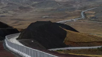 L'image montre un paysage vaste et aride, avec des collines et des montagnes en arrière-plan. Au premier plan, il y a un mur de béton qui serpente le long du sol, suivi d'une route qui s'étend dans la vallée. Le terrain est principalement composé de terre brune et de végétation clairsemée, avec des déclivités et des pentes. L'ambiance générale semble désertique et isolée.