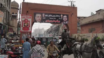 L'image montre une rue animée d'une ville avec un grand panneau publicitaire suspendu au-dessus. Ce panneau affiche des portraits de candidats politiques, accompagnés d'un message de campagne. Au premier plan, on voit des véhicules variés, y compris des charrettes tirées par des chevaux, des motos et des bicyclettes, illustrant l'activité de la vie urbaine. En arrière-plan, on peut apercevoir des bâtiments typiques et une ambiance dynamique, avec des personnes se déplaçant dans la rue.
