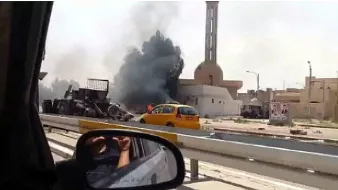 L'image montre une scène d'explosion ou d'incendie, avec de la fumée noire s'échappant d'un bâtiment en arrière-plan. On aperçoit également un véhicule jaune sur la route, tandis qu'une personne semble prendre la photo depuis l'intérieur d'une voiture. Des débris sont visibles près de l'endroit de l'incident, qui pourrait être lié à un conflit. La scène évoque une atmosphère de chaos et de tension.