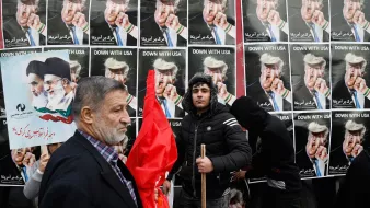 L'image montre une manifestation avec des affiches en arrière-plan affichant des personnages aux visages provocateurs, probablement des figures politiques, portant des coiffures spécifiques. Des gens tiennent des pancartes et des drapeaux, tandis qu'un homme sur le devant semble exprimer un message clair de désaccord ou de protestation. L'ensemble de la scène dégage une ambiance de contestation, soulignant des tensions politiques.