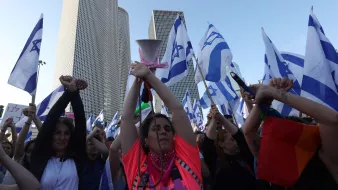 L'image montre une foule de manifestants tenant des drapeaux israéliens. Au centre, une femme portant un haut fluorescent rose élève les bras, tenant un mégaphone. Les manifestants semblent être engagés et passionnés, réunis devant des bâtiments modernes qui dominent l'arrière-plan. L'atmosphère semble être festive et revendicative.