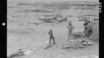 L'image montre une scène archéologique dans un paysage désertique. Plusieurs hommes, habillés de manière décontractée et portant des chapeaux, sont visibles. Ils semblent discuter ou examiner des fragments de pierres et des structures en ruines au sol. En arrière-plan, on peut voir des sites de fouille avec des vestiges qui suggèrent une activité archéologique en cours. L'environnement est aride, et le ciel paraît clair, évoquant une ambiance de travail sur un site ancien.