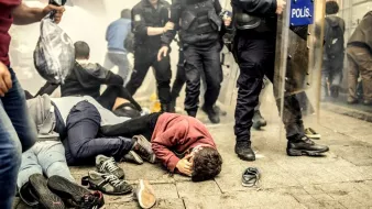 L'image montre une scène de manifestation ou d'émeute. On peut voir plusieurs personnes au sol, certaines semblant blessées ou cernées par des forces de police. Des policiers en tenue anti-émeute avancent dans une atmosphère de chaos, avec des gaz lacrymogènes obscurcissant l'air. L'environnement paraît tendu, témoignant d'un conflit entre manifestants et autorités.