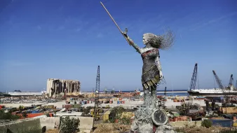 L'image présente une sculpture en métal d'une femme levant un bâton, symbolisant probablement la force et la résilience. Elle se tient sur une plateforme, surplombant un paysage industriel ou portuaire, avec des conteneurs, des grues et des bâtiments en arrière-plan. On peut également apercevoir des vestiges d'un bâtiment détruit, suggérant une scène de reconstruction ou de renouveau après une catastrophe. Le ciel est dégagé et ensoleillé, contrastant avec l'environnement chaotique du port.