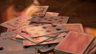L'image montre un tas de cartes à jouer éparpillées sur une surface. On peut voir différents types de cartes, y compris des cartes de cœur, de carreau, de trèfle et de pique. Certaines cartes semblent retournées, tandis que d'autres sont face visible, montrant leurs motifs et valeurs. L'éclairage apporte une ambiance chaleureuse et met en valeur la texture des cartes.