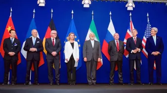 L'image montre un groupe de personnalités politiques posant ensemble devant un mur de drapeaux. On peut voir des représentants de plusieurs pays avec des drapeaux de la Chine, de l'Allemagne, de l'Union européenne, de l'Iran, de la Russie, du Royaume-Uni et des États-Unis en arrière-plan. Les personnes semblent discuter d'un sujet important et portent des costumes formels. L'ambiance est sérieuse, correspondant à un événement diplomatique.