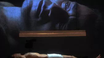 L'image montre un homme allongé sur une surface, partiellement nu, avec une serviette couvrant son corps. Au-dessus de lui, une projection de son visage est affichée sur un grand écran, créant un effet visuel puissant. La lumière provient d'une source suspendue, ajoutant une atmosphère dramatique à la scène. Les détails du visage projeté sont clairement visibles, tandis que l'homme au sol semble dans un état de vulnérabilité. L'ensemble évoque des thèmes de réflexion, d'introspection ou de méditation.