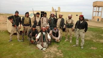 L'image montre un groupe de personnes en tenue militaire, armées et posant devant un véhicule. Ils semblent être sur un terrain en extérieur, peut-être dans un environnement désertique ou rural. Les hommes affichent diverses expressions et postures, certains sont debout tandis que d'autres sont assis. Leurs vêtements et accessoires suggèrent qu'ils font partie d'une unité militaire ou paramilitaire. En arrière-plan, on aperçoit un bâtiment et une tour d'eau.