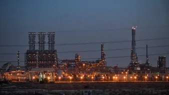 L'image montre une grande installation industrielle, probablement une raffinerie ou une usine de traitement, éclairée par de nombreuses lumières à la tombée de la nuit. On peut voir de hautes structures vertes et des cheminées qui s'élèvent dans le ciel. L'environnement autour semble désertique, avec peu de végétation visible. Les lignes électriques traversent le premier plan, ajoutant à l'atmosphère de l'image. Les lumières orange et blanches des installations créent un contraste avec le ciel sombre.