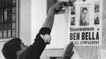 L'image montre un homme en train de coller une affiche sur un mur. L'affiche annonce un message de bienvenue à Ben Bella et ses compagnons, tout en affichant plusieurs portraits. L'homme semble concentré et utilise un outil pour bien lisser l'affiche contre la surface. L'environnement urbain suggère une période historique, probablement liée à un événement important. Les détails architecturaux et la tenue de l'homme donnent une impression de contexte social et culturel spécifique.