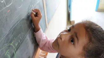 L'image montre une jeune fille penchée vers un tableau noir. Elle semble concentrée alors qu'elle écrit ou dessine quelque chose avec une craie. On peut voir des lettres ou des symboles tracés sur le tableau. La lumière entre par une fenêtre visible en arrière-plan, créant une ambiance chaleureuse. La fille porte un sweat à capuche rose et a les cheveux bouclés.