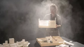 L'image montre une personne tenant une maquette d'un bâtiment, dans un environnement brumeux. Le fond est sombre, ce qui met en valeur la maquette et la silhouette de la personne. Sur la table, il y a d'autres maquettes et éléments architecturaux, suggérant une scène de travail ou de présentation liée à l'architecture. La lumière semble filtrer à travers la brume, créant une ambiance mystérieuse et artistique.