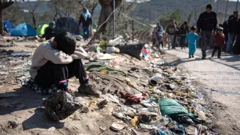L'image montre un jeune enfant assis au sol, semblant triste ou désemparé. Il est entouré de déchets et de débris, ce qui souligne une situation de pauvreté et de désespoir. À l'arrière-plan, on peut voir d'autres personnes marchant, probablement des familles, dans un environnement difficile. Des tentes sont visibles, indiquant un campement temporaire. L'ensemble de la scène évoque une réalité difficile et poignante.