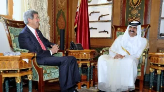 L'image montre deux hommes assis dans un bureau élégant, décoré avec des motifs riches et des couleurs vives. L'un des hommes porte un costume formel et sourit, tandis que l'autre est vêtu d'une tenue traditionnelle blanche, avec un chapeau noir. Ils semblent être en train de discuter dans une atmosphère amicale. En arrière-plan, on peut voir des objets décoratifs et des armes anciennes accrochées au mur, ajoutant à l'ambiance opulente de la pièce.