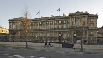 L'image montre un bâtiment imposant avec une architecture classique, situé dans un espace urbain. On peut voir des drapeaux français flottant au sommet, et le bâtiment est entouré d'une grille. La façade est riche en détails, avec des colonnes et des ornements décoratifs. Il semble être un édifice gouvernemental ou historique, situé probablement dans une grande ville française. Des arbres sont visibles sur les côtés, et une route passe devant le bâtiment.
