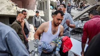 L'image montre une scène de désastre, probablement suite à un bombardement ou un tremblement de terre. On voit des décombres partout, avec des bâtiments endommagés. Un homme porte un enfant, probablement blessé, et semble préoccupé. D'autres personnes autour semblent s'affairer, cherchant à aider ou à évacuer les victimes. L'atmosphère est sombre et tragique, reflétant la détresse des personnes présentes.