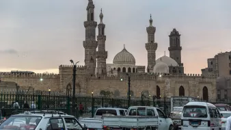 L'image montre un paysage urbain avec des bâtiments emblématiques, comprenant des mosquées avec des minarets élancés et des dômes caractéristiques. Au premier plan, on peut voir des véhicules traversant une route animée, entourés d'une clôture verte. L'éclairage doux de la fin de journée ajoute une atmosphère chaleureuse à la scène, tandis que le ciel présente des teintes nuancées, suggérant le coucher du soleil. L'architecture évoque un style traditionnel, typique de certaines grandes villes historiques.