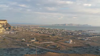 L'image montre un paysage côtier avec une vue panoramique. On peut voir une ville située au bord de la mer, entourée de collines et de montagnes à l'horizon. La mer est calme, et des structures côtières, comme des docks ou des bateaux, sont visibles. Le ciel est partiellement nuageux, ce qui donne une belle lumière à la scène. On observe également des zones désertiques ou semi-désertiques, avec des routes qui serpentent à travers le paysage.