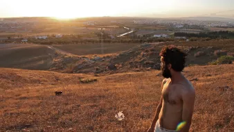 L'image montre un homme debout sur une colline, regardant vers un paysage vallonné au coucher du soleil. Il a les cheveux et la barbe bruns, et porte un t-shirt. Le terrain autour de lui est sec et couvert d'herbes hautes. En arrière-plan, on peut voir un petit village et des champs qui s'étendent à perte de vue, créant un panorama paisible et naturel. La lumière du soleil met en valeur les couleurs dorées de la scène.