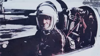 L'image montre un pilote assis dans le cockpit d'un avion ou d'un vaisseau spatial. Il porte un casque de protection et une combinaison, ce qui indique qu'il est probablement en préparation pour un vol. Le décor autour de lui semble être un environnement aéroporté ou une piste de lancement. L'ambiance de l'image évoque la technologie et l'exploration aérienne ou spatiale.