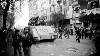L'image montre une scène de manifestation urbaine, avec des gens dans une rue animée. Au premier plan, plusieurs manifestants marchent, certains levant les bras en l'air. À l'arrière-plan, un tank est visible, portant des inscriptions. La scène est en noir et blanc, ce qui intensifie l'atmosphère dramatique et chargée d'émotion. Les bâtiments environnants et les arbres ajoutent à l'environnement urbain. L'ensemble dégage un sentiment de tension et de mouvement.