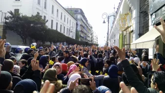 L'image montre une grande foule de personnes rassemblées dans une rue, levant les mains en signe de protestation ou de solidarité. On peut voir des bâtiments architecturaux en arrière-plan, ainsi que quelques agents de police en uniforme. Les participants semblent très engagés dans leur manifestation, et l'atmosphère est chargée d'énergie collective. Les vêtements des manifestants varient, certains portant des écharpes ou des bonnets en fonction de la saison.