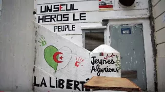 L'image montre un mur peint avec plusieurs graffitis. On y trouve des mots et des phrases exprimant des messages politiques et sociaux. Un dessin représentant un cœur dans les couleurs du drapeau algérien (rouge et vert) est visible. Il y a aussi une inscription en arabe, ainsi que des mots tels que "Jeunies Algériens" et "LA LIBERTÉ". Le fond suggère un lieu qui pourrait être un espace public, témoignant d'une expression artistique et militante.
