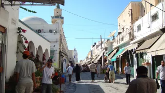 L'image montre une rue animée d'une ville typique, avec des bâtiments de style méditerranéen. On peut voir des personnes marchant le long de la rue, parmi lesquelles des hommes habillés en vêtements traditionnels. Des pots de fleurs et des végétaux décorent l'entrée de certains bâtiments, tandis que des ombrelles ou des auvents semblent offrir de l'ombre aux passants. Le ciel est dégagé et ensoleillé, créant une ambiance agréable.