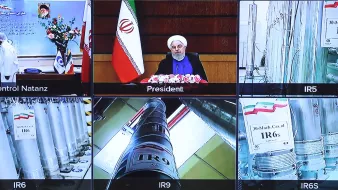 L'image montre une composition de plusieurs écrans de télévision. Au centre, on voit un homme, probablement un président, assis devant un bureau avec des fleurs, entouré de drapeaux iraniens. Sur les autres écrans, il y a des images d'installations ou d'équipements liés à la technologie nucléaire, avec des tubes et des étiquettes indiquant différentes séries de centrifugeuses nucléaires. Il y a aussi des symboles et des drapeaux qui décrivent les différentes installations mentionnées. L'ensemble semble se rapporter à un discours ou une présentation sur le programme nucléaire de l'Iran.