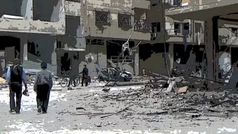 L'image montre une scène de destruction dans une zone urbaine. On y voit des bâtiments endommagés, avec des débris éparpillés sur le sol. Deux personnes marchent au premier plan, tandis que d'autres personnes sont visibles au loin, certaines semblent conduire des vélos. L'ambiance générale est triste et chaotique, illustrant les conséquences dévastatrices d'un conflit.